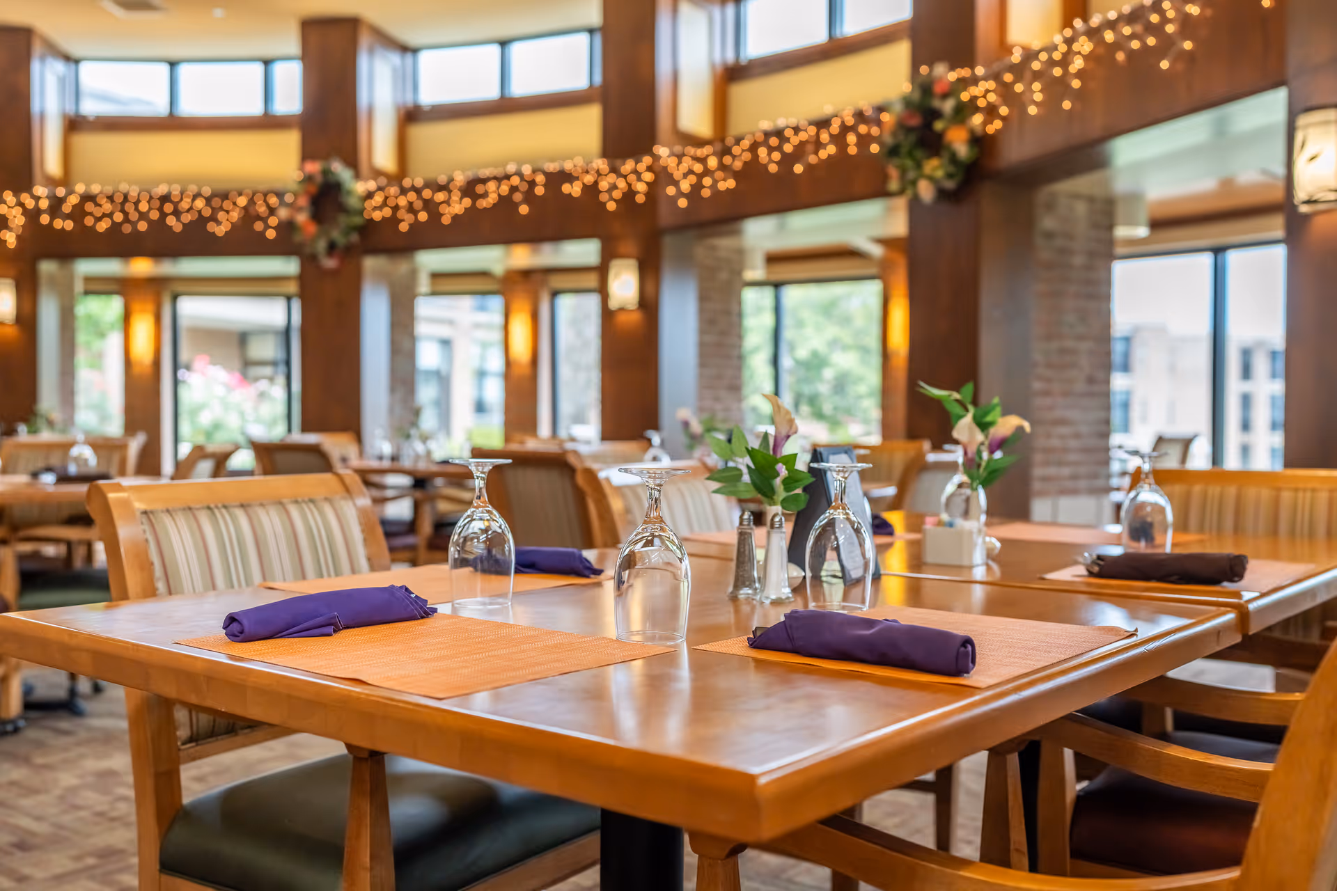 A well-lit dining room with wooden tables set with orange placemats, purple napkins, upside-down wine glasses, small flower arrangements, and salt and pepper shakers. The room features large windows, wooden columns, and string lights hanging along the upper walls.