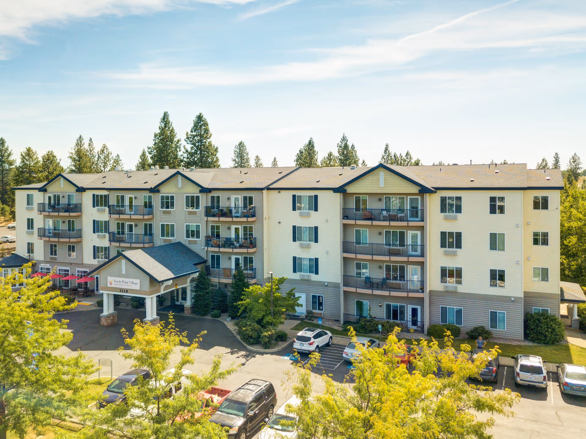 Exterior view of a multi-story senior living facility named North Point Village, surrounded by trees and a parking lot with several cars parked. The building has balconies and a covered entrance.