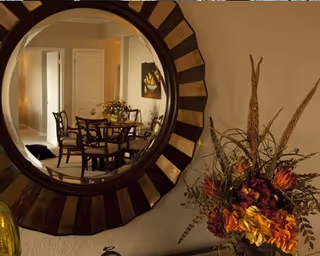 A round decorative mirror on a wall reflecting a dining area with a wooden table and chairs, a vase with flowers on the table, and a closed door in the background. Next to the mirror is a floral arrangement with orange and yellow flowers and greenery.