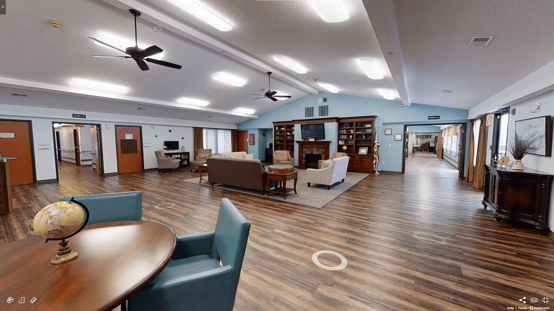 Spacious communal living room in a senior living facility with seating arranged around a fireplace, wood floors, and a small table with a globe in the foreground.