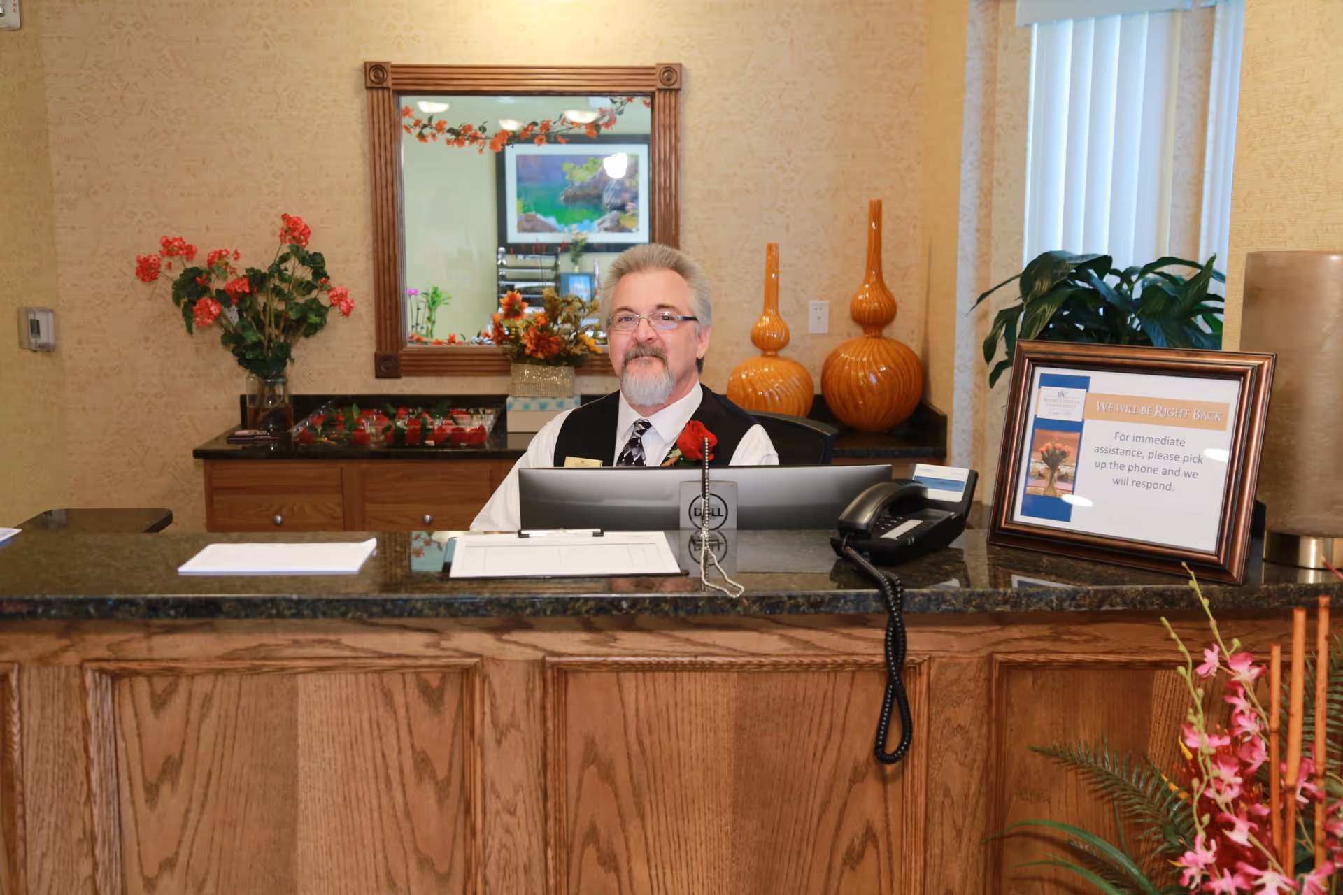 A man with gray hair and a beard, wearing glasses, a white shirt, black vest, and tie, sits behind a wooden reception desk with a granite countertop. The desk has a computer monitor, a telephone, and a framed sign that reads 'We will be right back. For immediate assistance, please pick up the phone and we will respond.' Behind the man, there is a wall mirror, decorative orange vases, and a plant with red flowers. The room has beige wallpaper and vertical blinds on the window.