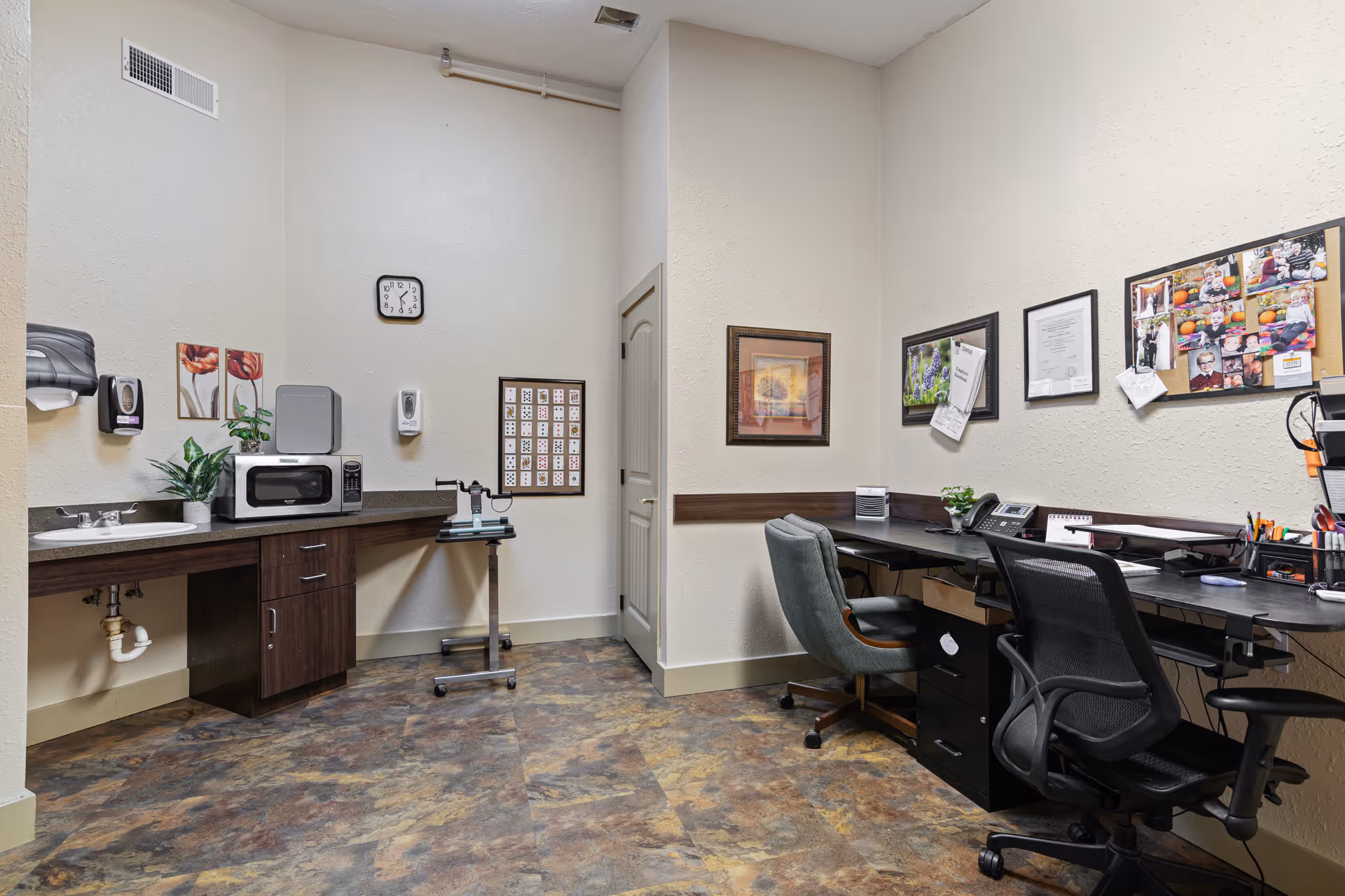 Small staff office area with desks, chairs, a sink, microwave and bulletin boards in a nursing facility.