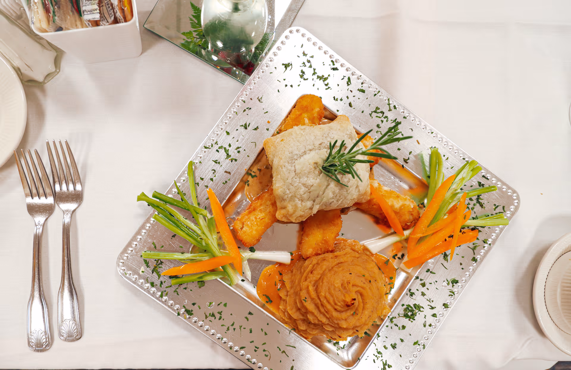 A plated meal on a silver square plate featuring a biscuit topped with a sprig of rosemary, several fried items, a swirl of mashed sweet potatoes, and garnished with sliced carrots and green onions. The plate is on a white tablecloth with two forks on the left and a small container of condiments in the top left corner.