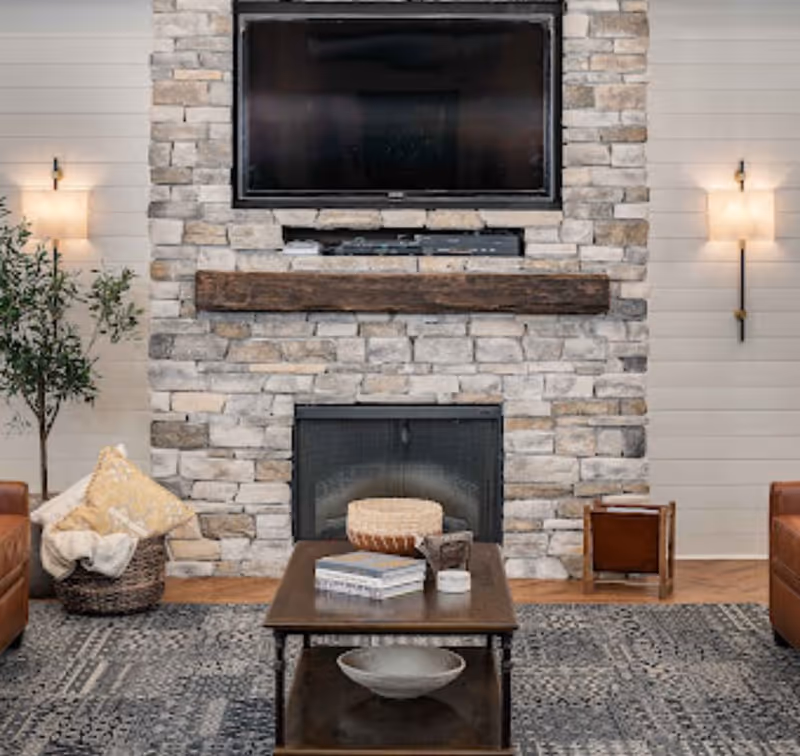 Cozy living room with a stone fireplace, mounted TV, wooden mantel, coffee table, rug and leather seating.