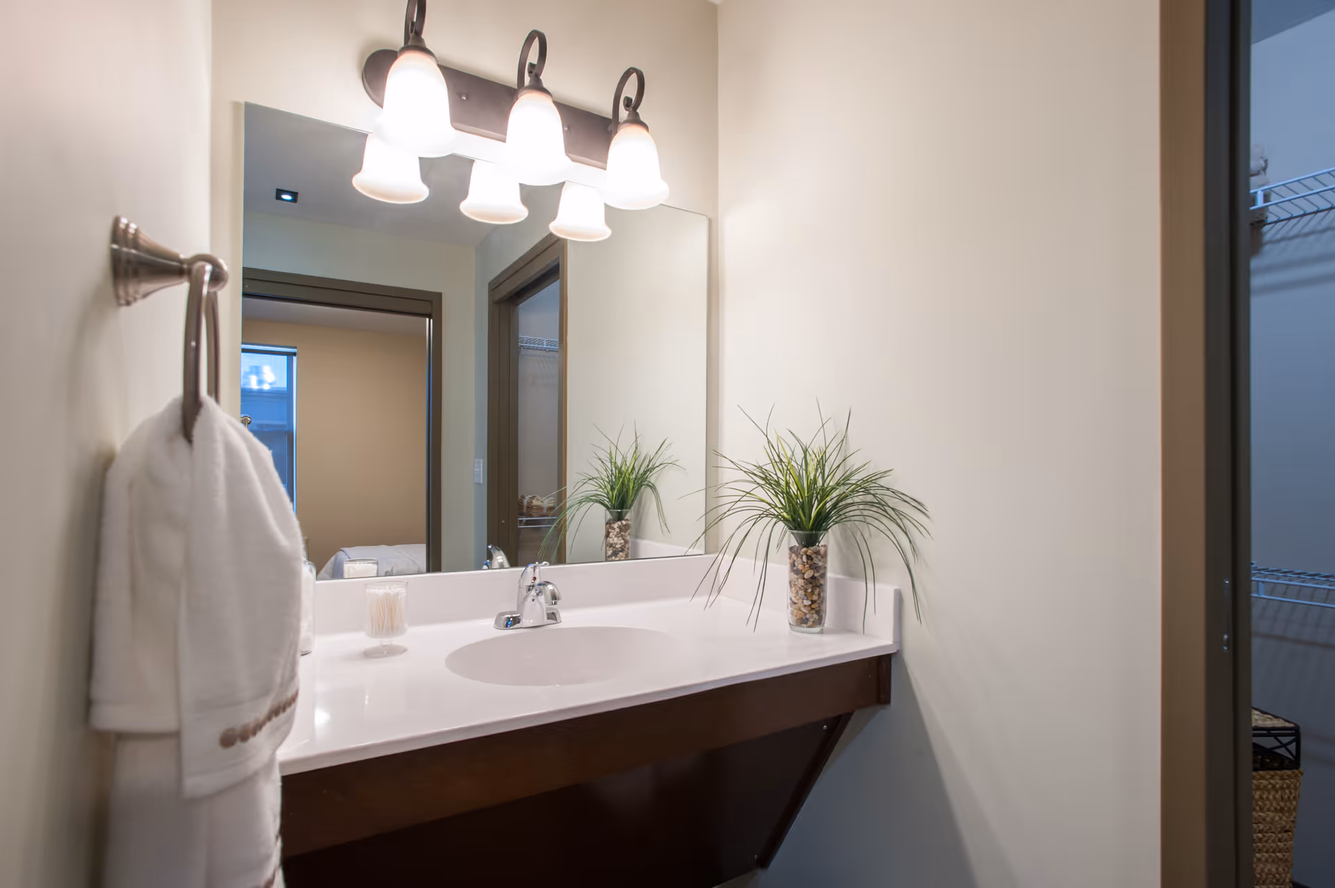 A clean bathroom sink area with a white countertop, a built-in sink, and a large mirror above. There is a three-light fixture mounted above the mirror. A white towel hangs on a towel ring on the left wall, and a small plant in a glass vase filled with stones is placed on the right side of the countertop. The reflection in the mirror shows a doorway and part of an adjacent room.