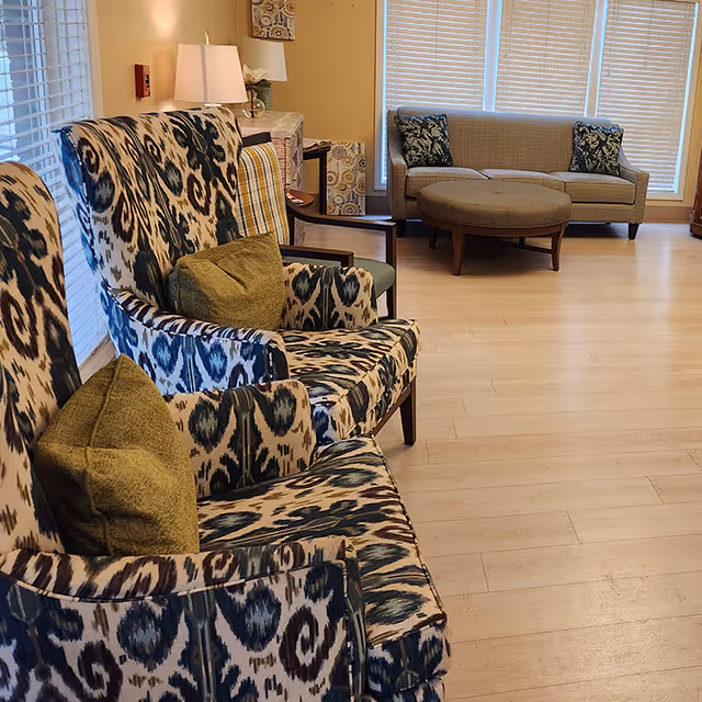 A cozy seating area in a living room with patterned armchairs featuring green cushions in the foreground and a beige sofa with two patterned pillows and a round ottoman in the background. The room has wooden flooring, large windows with blinds, and a lamp on a side table near the wall.