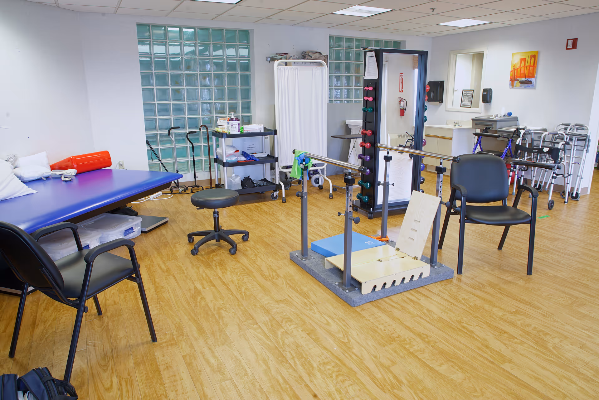 A physical therapy or rehabilitation room with wooden flooring, parallel bars for walking exercises, a black chair, a stool, a padded therapy table with pillows, various walkers and canes, a rack with colorful dumbbells, a mirror, and a privacy screen. The room has glass block windows and light-colored walls.