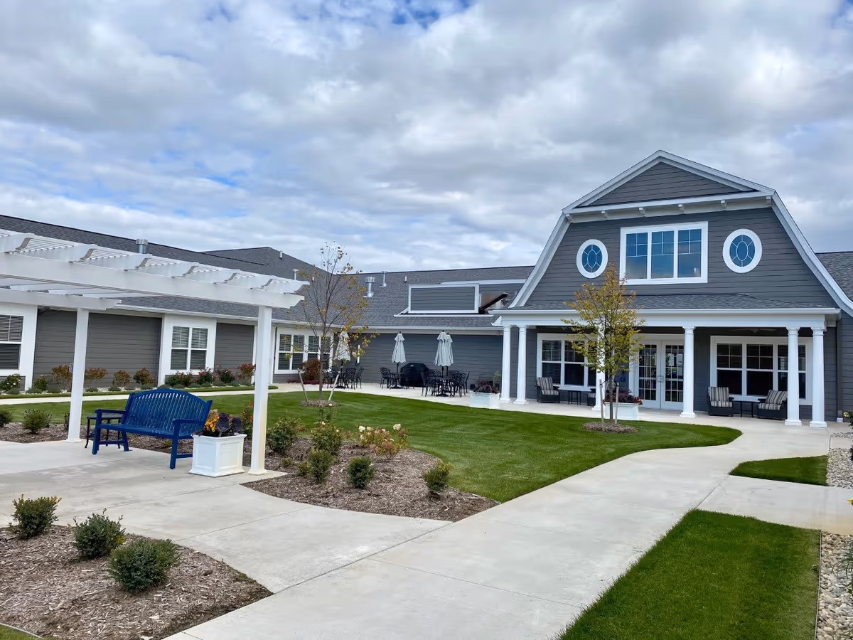 Outdoor courtyard area of Cascade Trails Senior Living featuring a gray building with white trim, a covered patio with seating, a white pergola with a blue bench underneath, landscaped garden beds, and a concrete walkway under a partly cloudy sky.