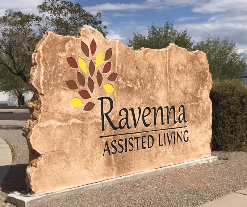 Large carved stone sign outdoors reading "Ravenna Assisted Living" with a stylized leaf logo.