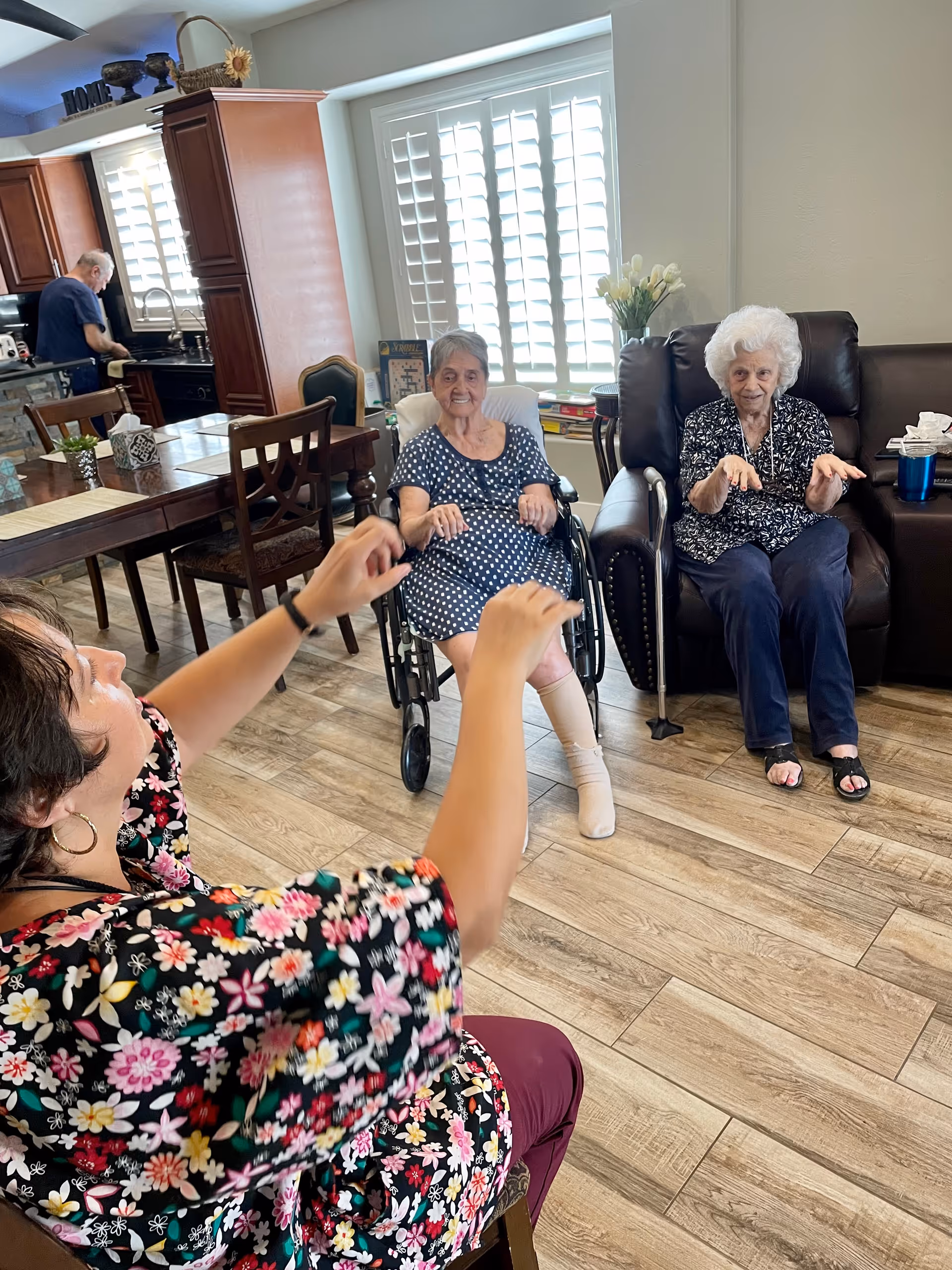 A group of elderly women and a caregiver engaging in a seated activity in a well-lit living area with wooden flooring. One elderly woman is in a wheelchair, another is seated in a recliner, and a caregiver with a floral top is in the foreground with raised hands. In the background, a man is working in the kitchen area.