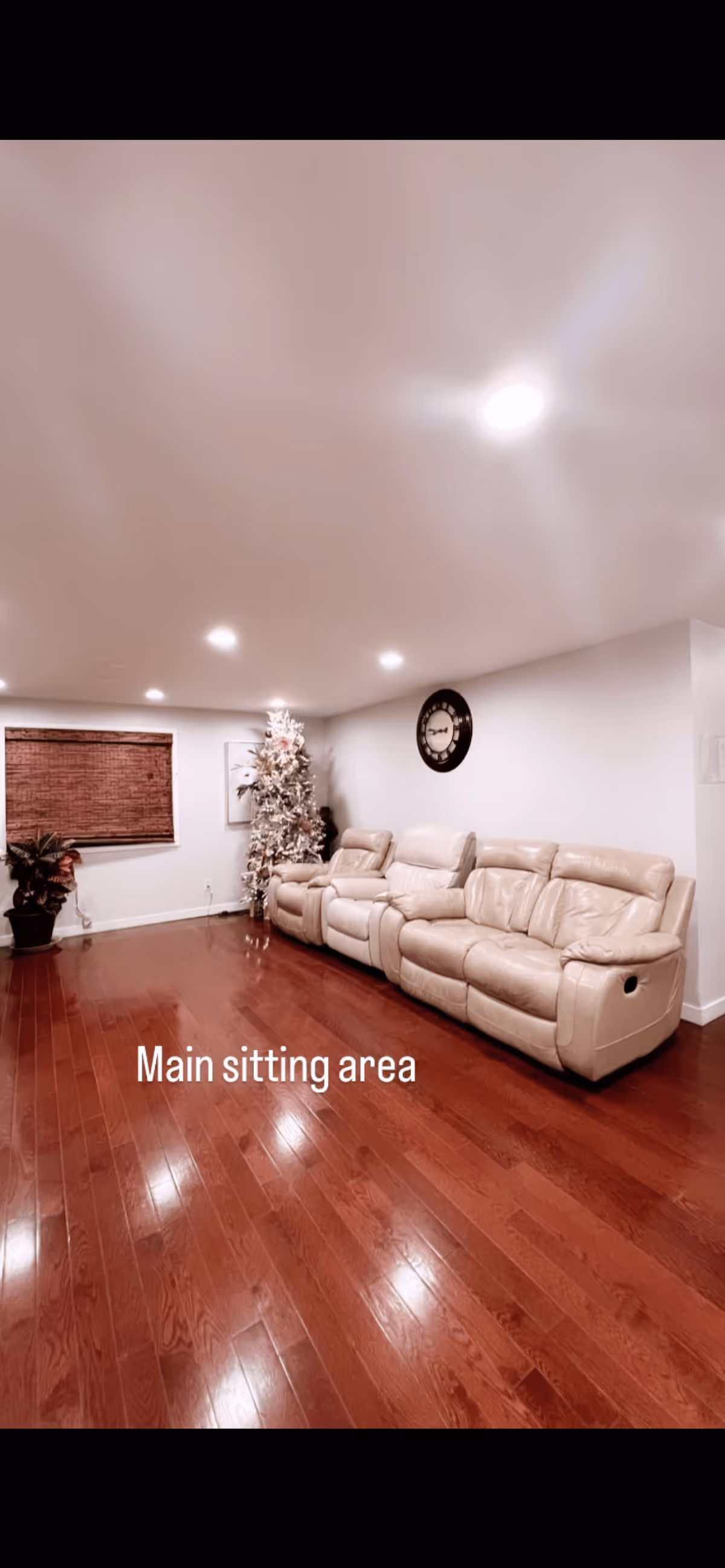 A spacious sitting area with polished wooden floors, a beige leather reclining sofa set against a white wall, a large round wall clock, a window with wooden blinds, a potted plant, and a decorated Christmas tree in the corner. The ceiling has recessed lighting.
