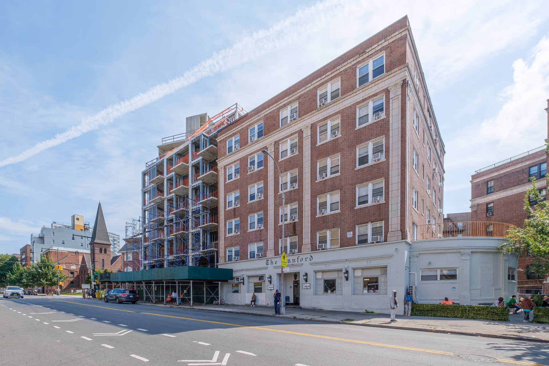 Exterior view of Sanford Manor, a multi-story brick building with white trim and multiple windows, located on a city street with a few people walking on the sidewalk and cars parked nearby. The building has a sign with the name Sanford and some scaffolding on one side indicating construction or renovation work.