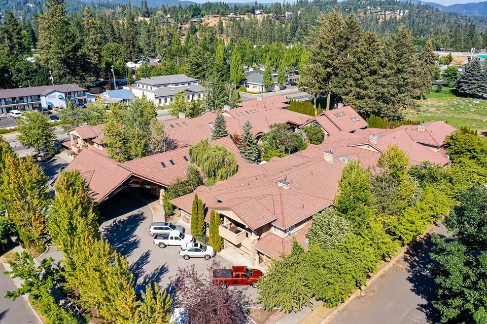 Aerial view of Courtyard at Coeur d'Alene senior living facility surrounded by trees and greenery, with multiple buildings featuring red roofs and a driveway with parked vehicles.