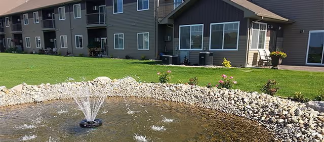 A landscaped lawn with a small decorative pond and fountain in front of a multi-unit senior living building.