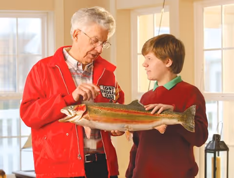 An elderly man wearing glasses and a red jacket is showing a large fish to a young boy in a red sweater inside a well-lit room with large windows.