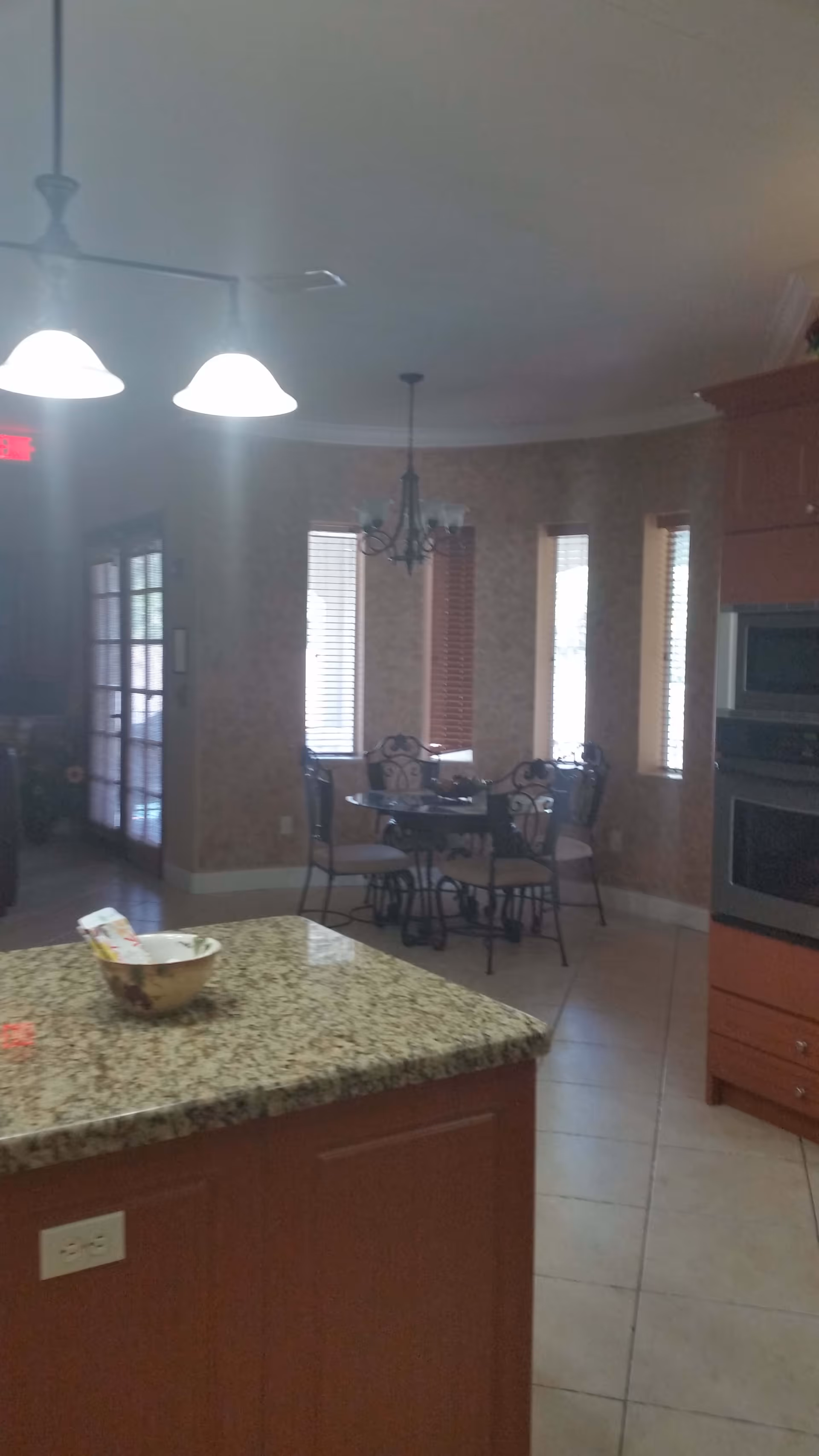 Interior view of a kitchen and dining area with a granite countertop island in the foreground, a round dining table with four chairs near windows with blinds, and built-in kitchen appliances on the right side.