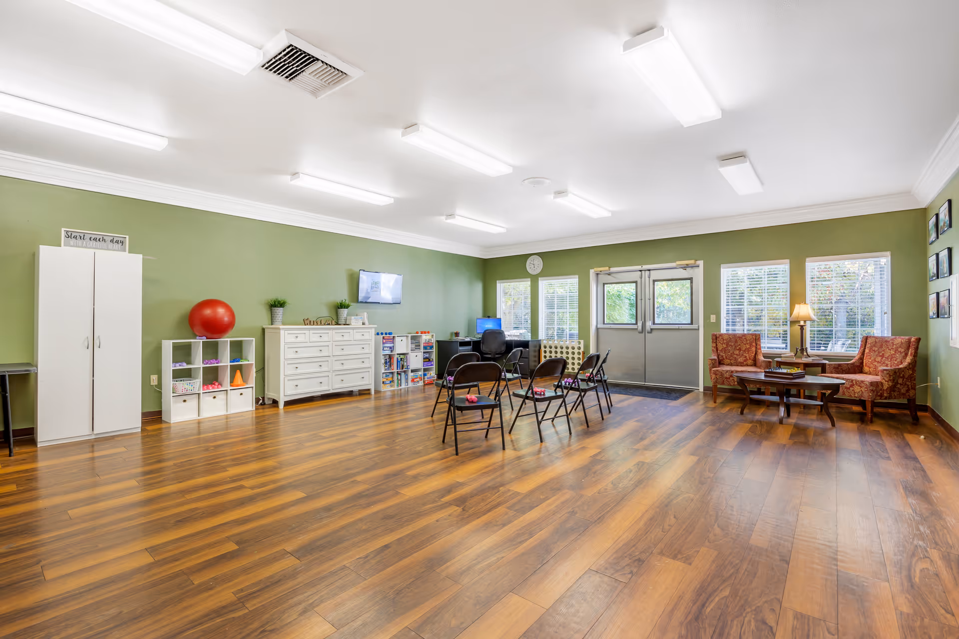 A spacious room with wooden flooring and green walls, featuring a small seating area with two patterned armchairs and a coffee table near windows, several black folding chairs arranged in a semi-circle, a white dresser, cubby shelves with various items, a red exercise ball, and a wall-mounted TV. The room is well-lit with ceiling lights and has double doors leading outside.