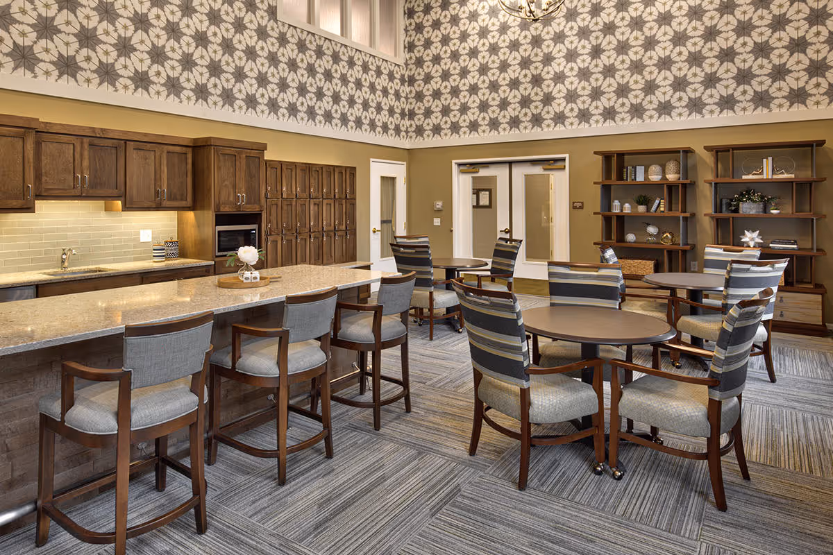 A spacious and well-lit communal dining area with a long kitchen island featuring four cushioned bar stools. The kitchen area has wooden cabinets, a microwave, and a sink with a tiled backsplash. There are several round tables with striped cushioned chairs arranged around the room. The walls have a decorative floral wallpaper near the ceiling, and there are two wooden bookshelves with decorative items. Double doors are visible in the background.