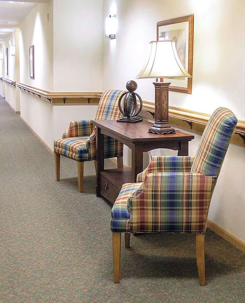 A hallway in a senior living facility with two plaid upholstered armchairs and a wooden table between them. The table holds a decorative lamp and a spherical sculpture. The walls are light-colored with wooden handrails and framed artwork, and the floor is carpeted.