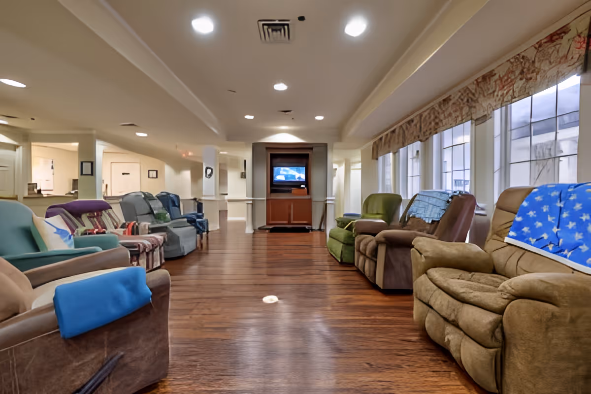 A long room with a row of various upholstered recliner chairs lined up on both sides facing each other. The room has wooden flooring, a ceiling with recessed lights, and large windows with patterned valances on the right side. At the far end of the room, there is a television set placed on a wooden cabinet.