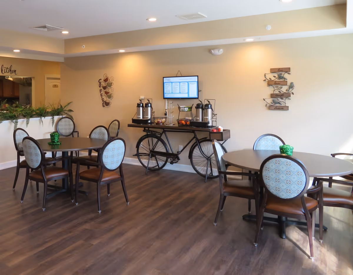 A dining area with two round tables, each surrounded by four chairs with patterned backs and brown seats. A decorative bicycle-shaped table against the wall holds coffee dispensers, fruit baskets, and other items. The wall has a mounted TV displaying a menu, a metal coffee cup wall decoration, and a wooden sign with the words 'live love laugh dream'. The floor is dark wood, and there is a partial view of a kitchen area to the left.