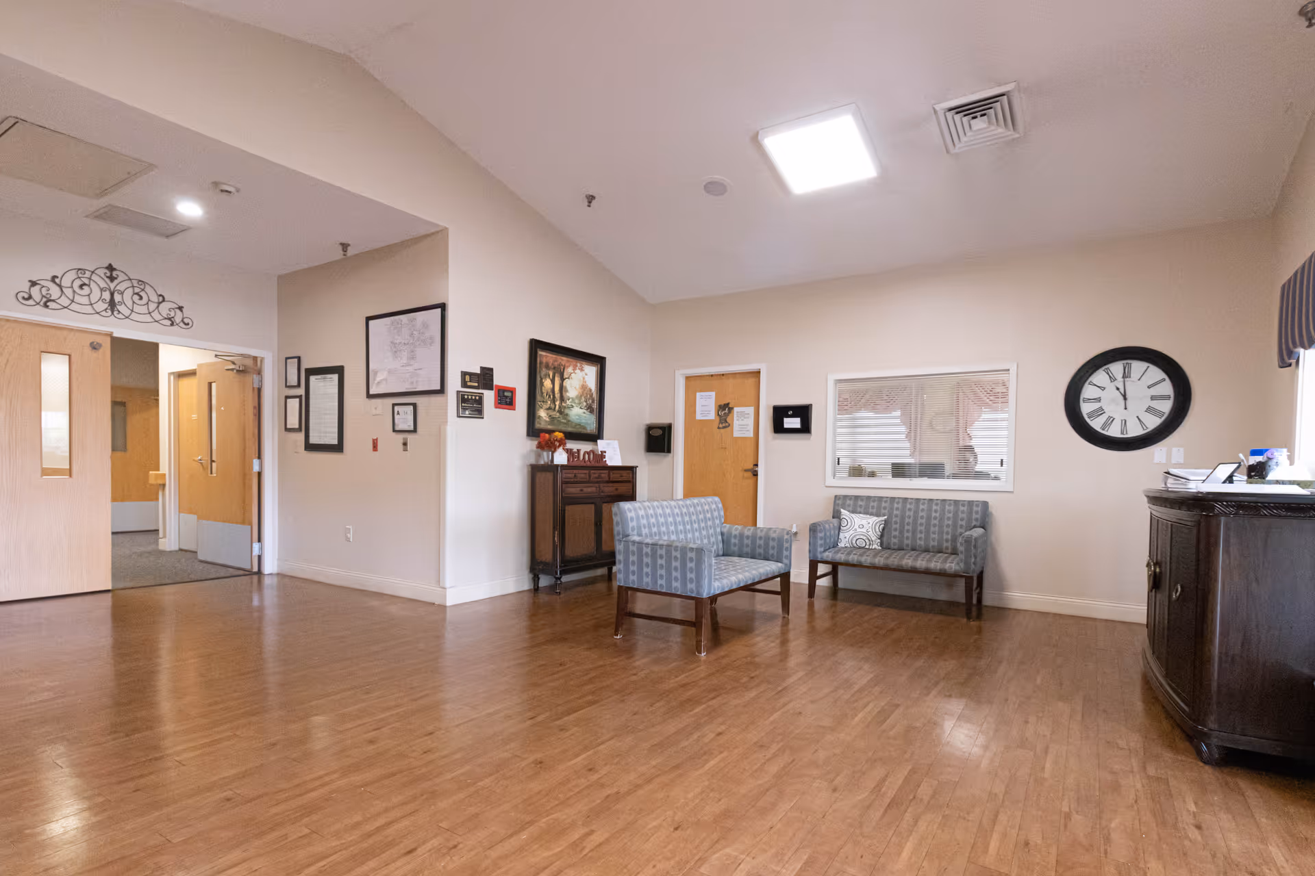 A bright senior living common room with wood floors, several upholstered chairs and a small sofa, a wall clock, and a reception counter.