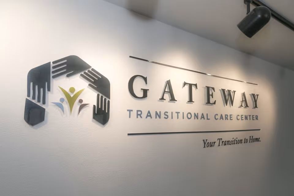 Wall sign inside a facility displaying the logo and name 'Gateway Transitional Care Center' with the tagline 'Your Transition to Home.' The logo features stylized hands forming a house shape around three abstract human figures.
