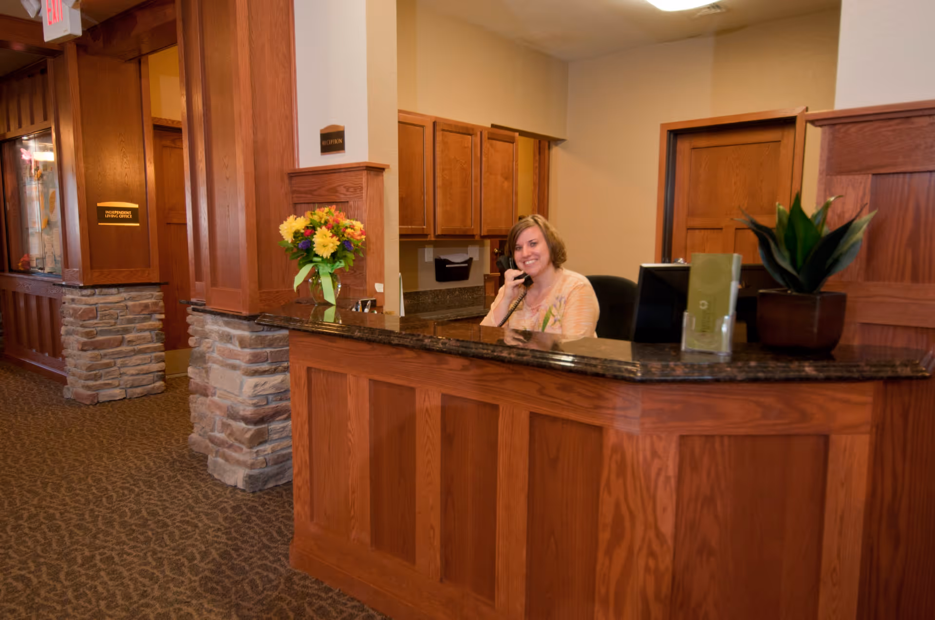 Reception area with a woman sitting behind a wooden desk, talking on the phone. The desk has a granite countertop with a vase of flowers and a potted plant. The background shows wooden cabinets and doors, with a sign labeled 'Reception'.