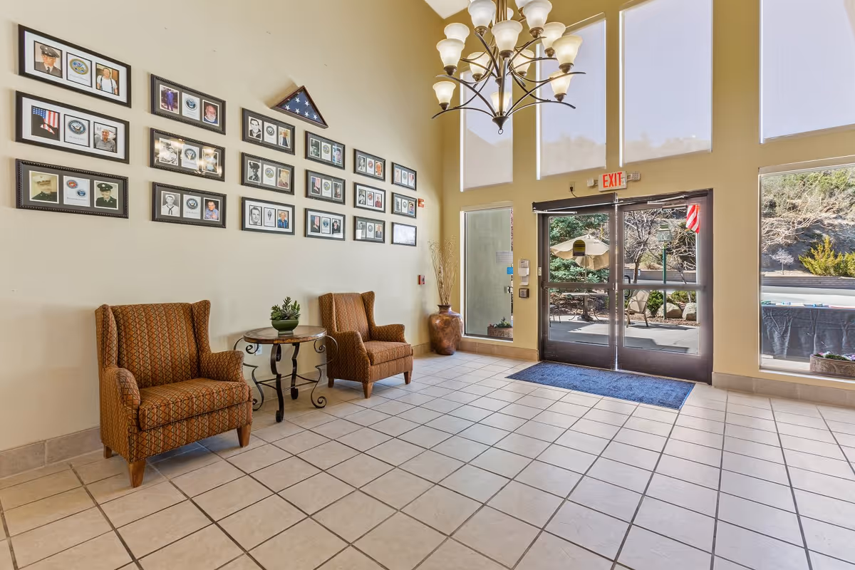 A bright lobby area with two patterned armchairs and a small round table between them. The wall behind the chairs is decorated with multiple framed photos and a folded American flag in a triangular display case. Large windows and glass doors let in natural light, showing an outdoor patio area with umbrellas and chairs. A chandelier hangs from the high ceiling.