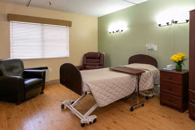 A simple, clean room with a single hospital-style bed covered with a beige quilted blanket. Next to the bed is a small wooden nightstand with a vase of yellow flowers and a few items on top. There is a brown recliner chair and a black leather armchair near a window with closed blinds. The walls are painted light green and beige, and the floor has a wood finish. Wall-mounted lights illuminate the room.