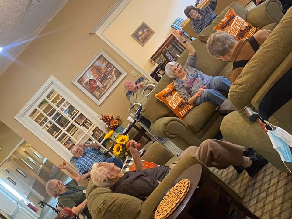 A group of elderly people sitting in a cozy living room with green couches and fall-themed pillows. They appear to be engaged in an activity, raising their hands and smiling. The room has warm beige walls, a painting, a table with a vase of sunflowers, and a plate of crackers on a side table.