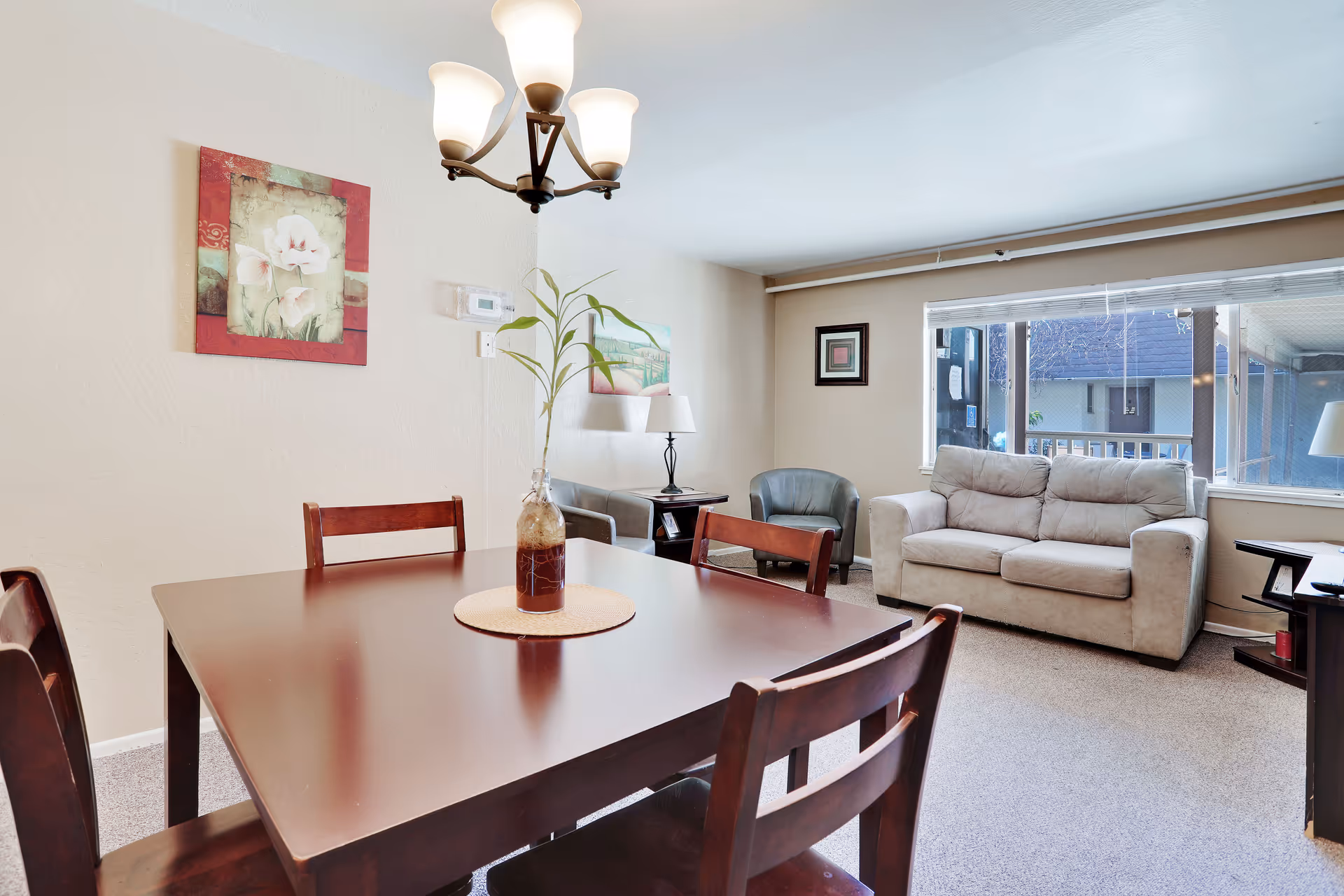 Sunny common room with a wooden dining table in the foreground, a sofa and armchairs by a large window, and wall art on the walls.