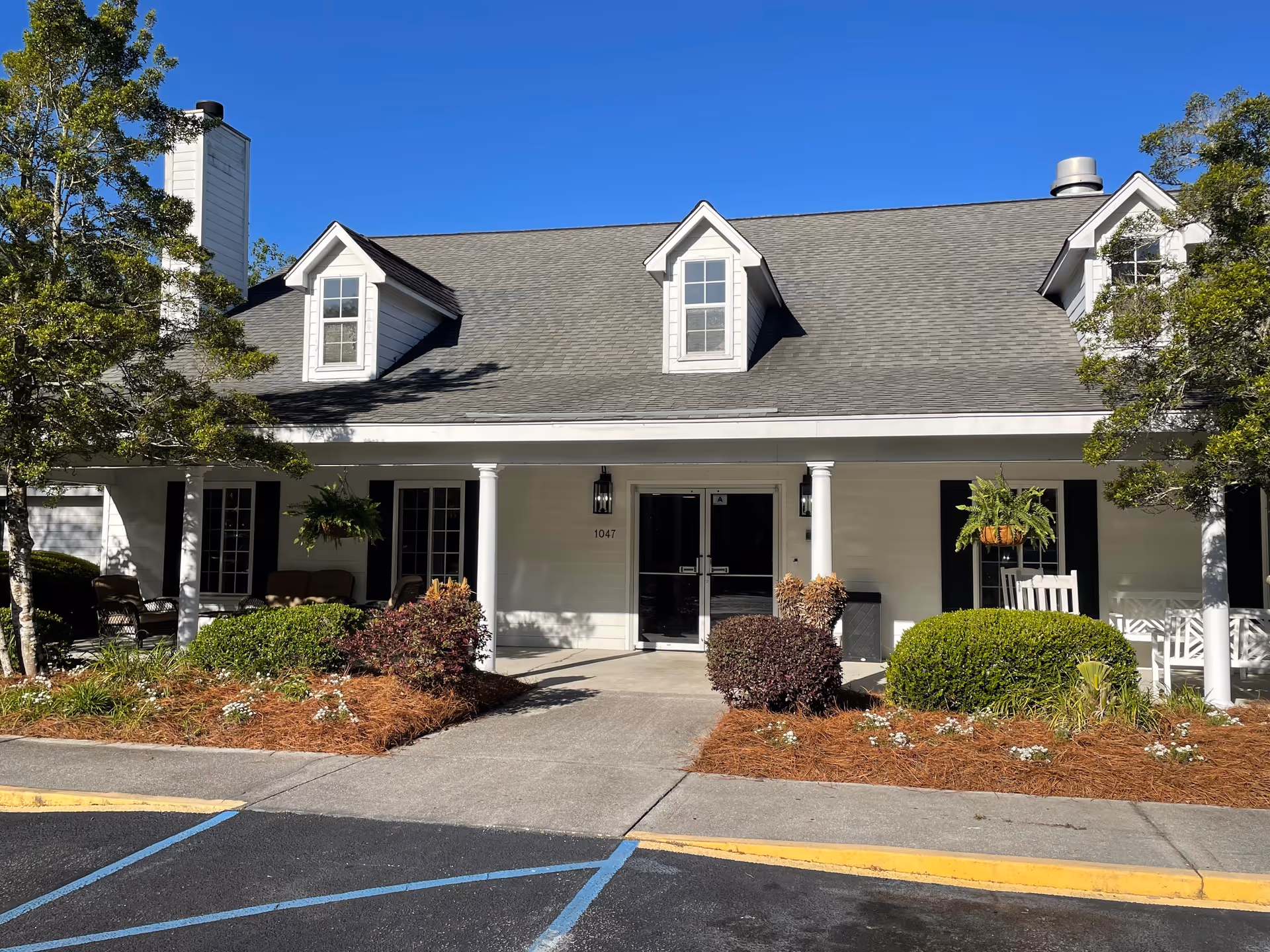 Front exterior view of a single-story building with a gray shingled roof, white siding, and three dormer windows. The entrance has double glass doors flanked by white columns and outdoor seating areas with chairs and hanging plants. There are landscaped bushes and flowers along the walkway leading to the entrance, and a clear blue sky overhead.