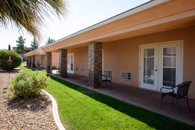 Outdoor view of a single-story building with a covered walkway supported by stone pillars. The building has beige walls and white-framed glass doors leading to individual rooms. There are black chairs placed along the walkway. The foreground features a well-maintained green lawn bordered by a curved edge with rocks and shrubs. A palm tree and other greenery are visible on the left side under a clear sky.