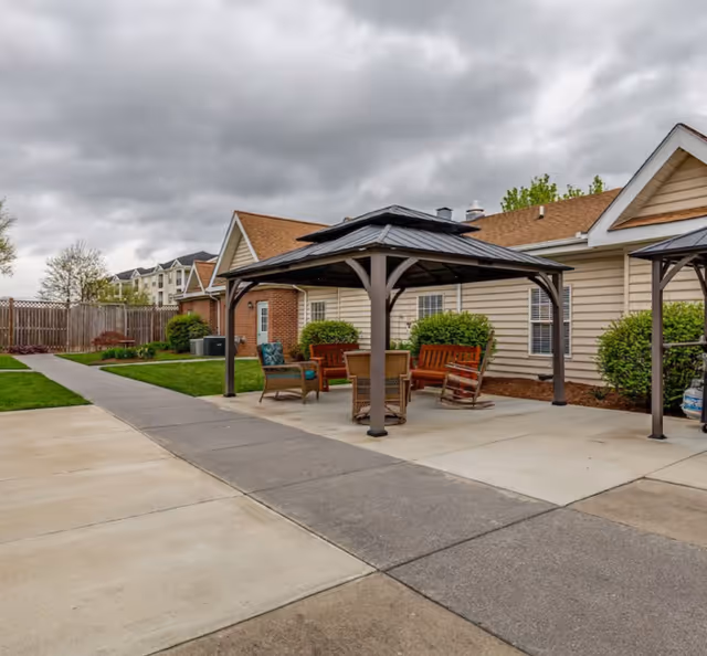 Outdoor patio area at Greenfield Senior Living at Cockeysville featuring a covered gazebo with wooden benches and chairs arranged around a table. The patio is adjacent to a single-story building with beige siding and a brown roof. There is a concrete walkway and green lawn with bushes and a wooden fence in the background under a cloudy sky.