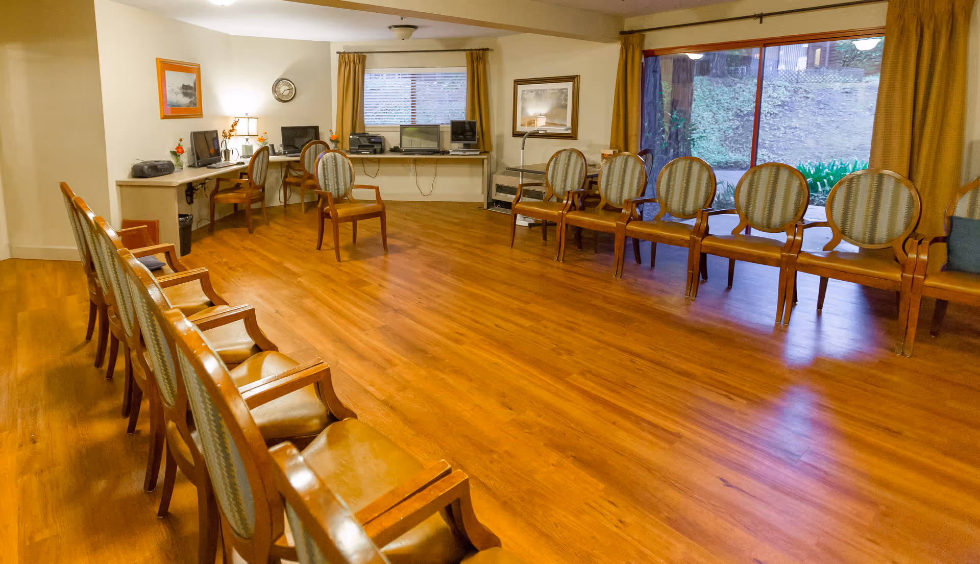 A spacious room with wooden flooring and two rows of wooden chairs with upholstered backs and seats arranged facing each other. The room has large windows with yellow curtains, a desk area with computers and lamps, framed pictures on the walls, and a clock above the desk.