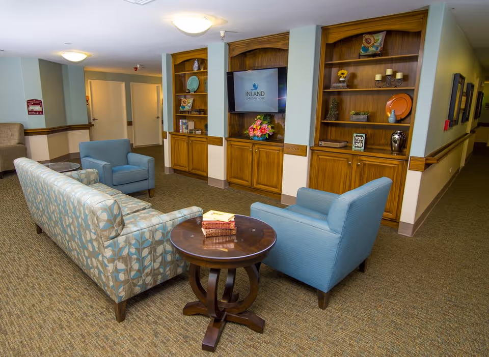 A cozy common area in a senior living facility with a patterned sofa, two blue armchairs, and a round wooden table with books on it. Behind the seating area are built-in wooden shelves and cabinets, with decorative items and a TV displaying the Inland Christian Home logo. The room has carpeted flooring and soft overhead lighting.