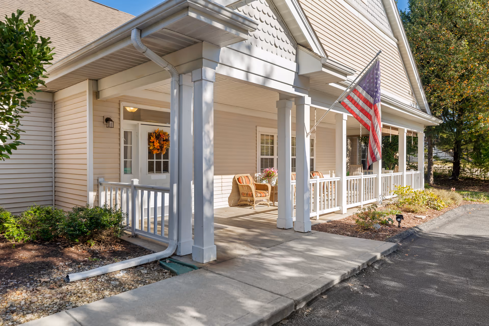Front porch of a building with beige siding, white columns, and a white railing. There are two wicker chairs with cushions and a small table with flowers on the porch. An American flag is mounted on one of the columns. The entrance door has a fall-themed wreath. There are shrubs and a paved walkway leading to the porch.