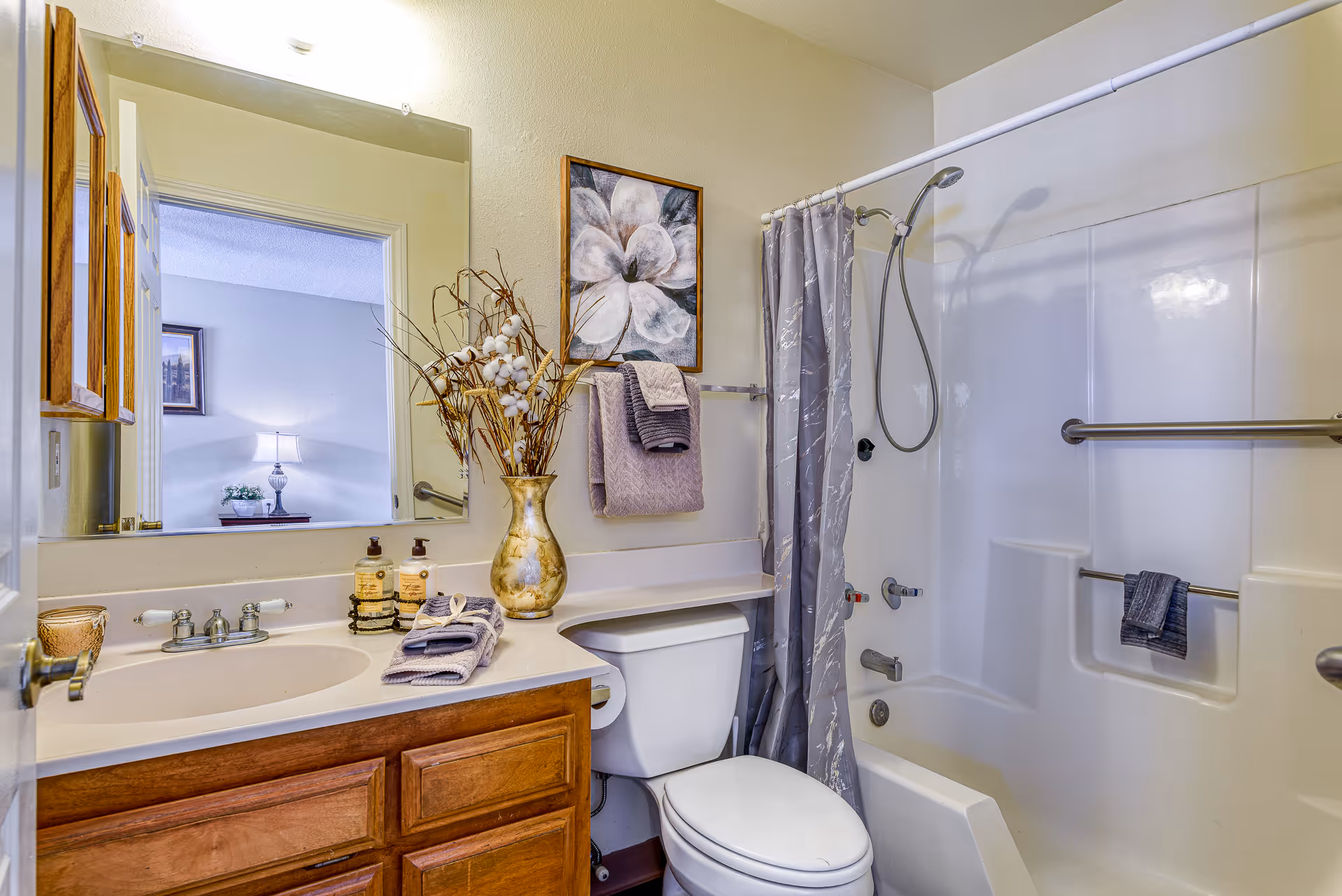 A bathroom with a wooden vanity cabinet, a white sink, and a large mirror above it. On the countertop, there is a decorative vase with dried branches and cotton, two bottles of soap or lotion, and neatly folded towels. A toilet is positioned next to the vanity. The bathtub has a showerhead, grab bars, and a gray shower curtain with a subtle pattern. A framed floral artwork hangs on the wall above the towel rack.