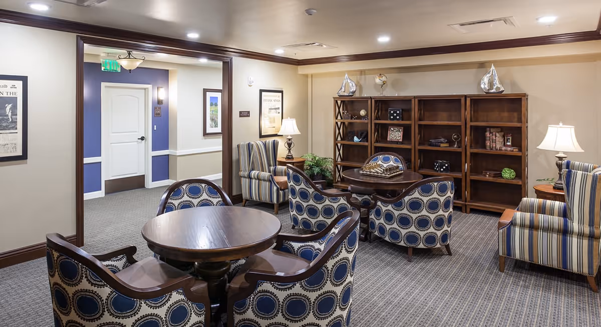 A cozy senior living common area with round wooden tables surrounded by upholstered chairs featuring blue and white circular patterns. There are two striped armchairs with side tables and lamps. A wooden bookshelf against the wall holds decorative items including model sailboats and globes. The room has beige walls with white trim and a carpeted floor. A hallway with a white door and framed pictures is visible in the background.