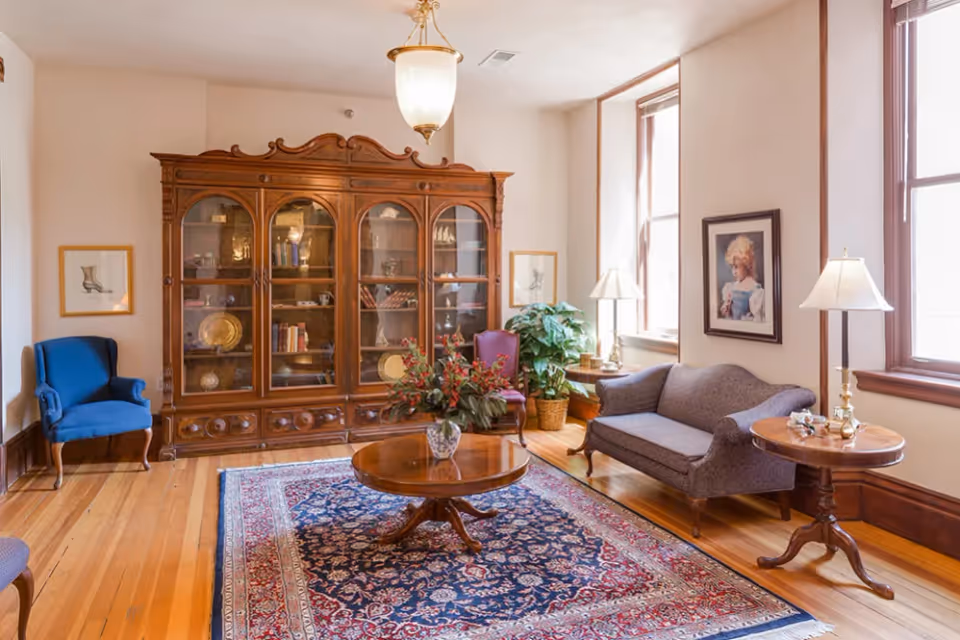 A cozy living room with wooden floors, a large ornate wooden cabinet with glass doors displaying decorative items and books, a round wooden coffee table with a floral arrangement, a blue armchair, a gray loveseat, two side tables with lamps, framed artwork on the walls, and large windows letting in natural light.