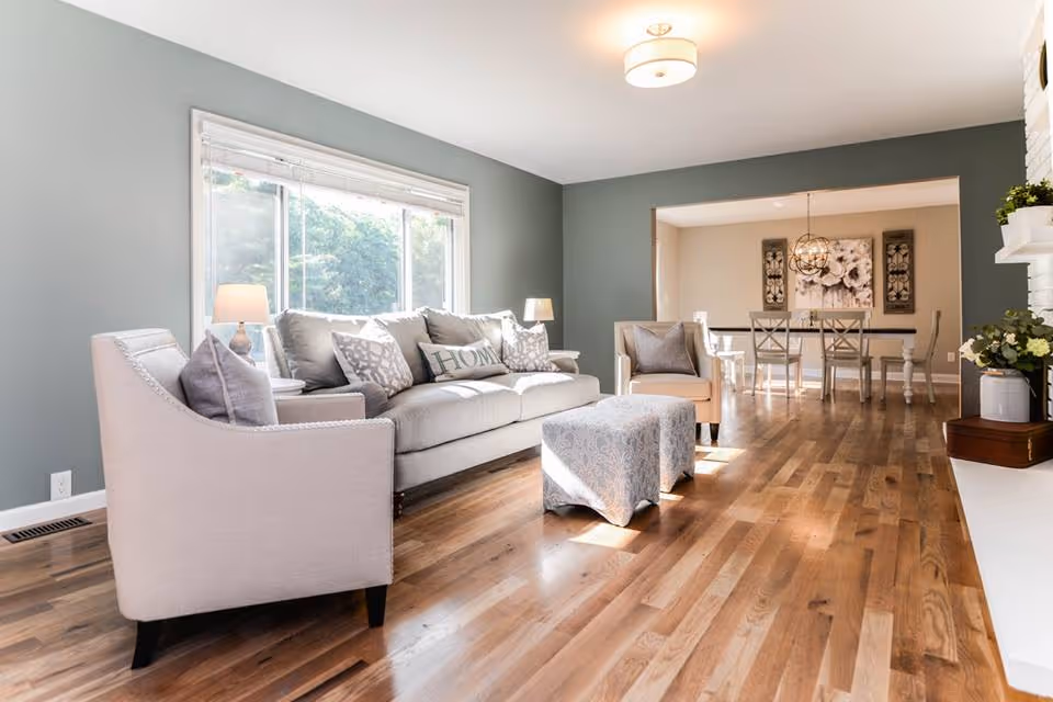 A bright and spacious living room with light gray walls and wooden flooring. The room features a light gray sofa with multiple cushions, including one with the word 'HOME' on it, two matching armchairs, and a patterned ottoman in the center. There are two table lamps on side tables next to the sofa. In the background, an open dining area is visible with a dining table, chairs, and wall art. The room is well-lit by natural light coming through a large window.