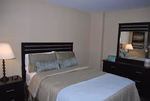 A neatly made bed with a dark wooden headboard, beige and blue bedding, and multiple pillows in a bedroom. To the left of the bed is a nightstand with a lamp and an alarm clock. To the right is a dark wooden dresser with a large mirror, a decorative vase, and a small basket.