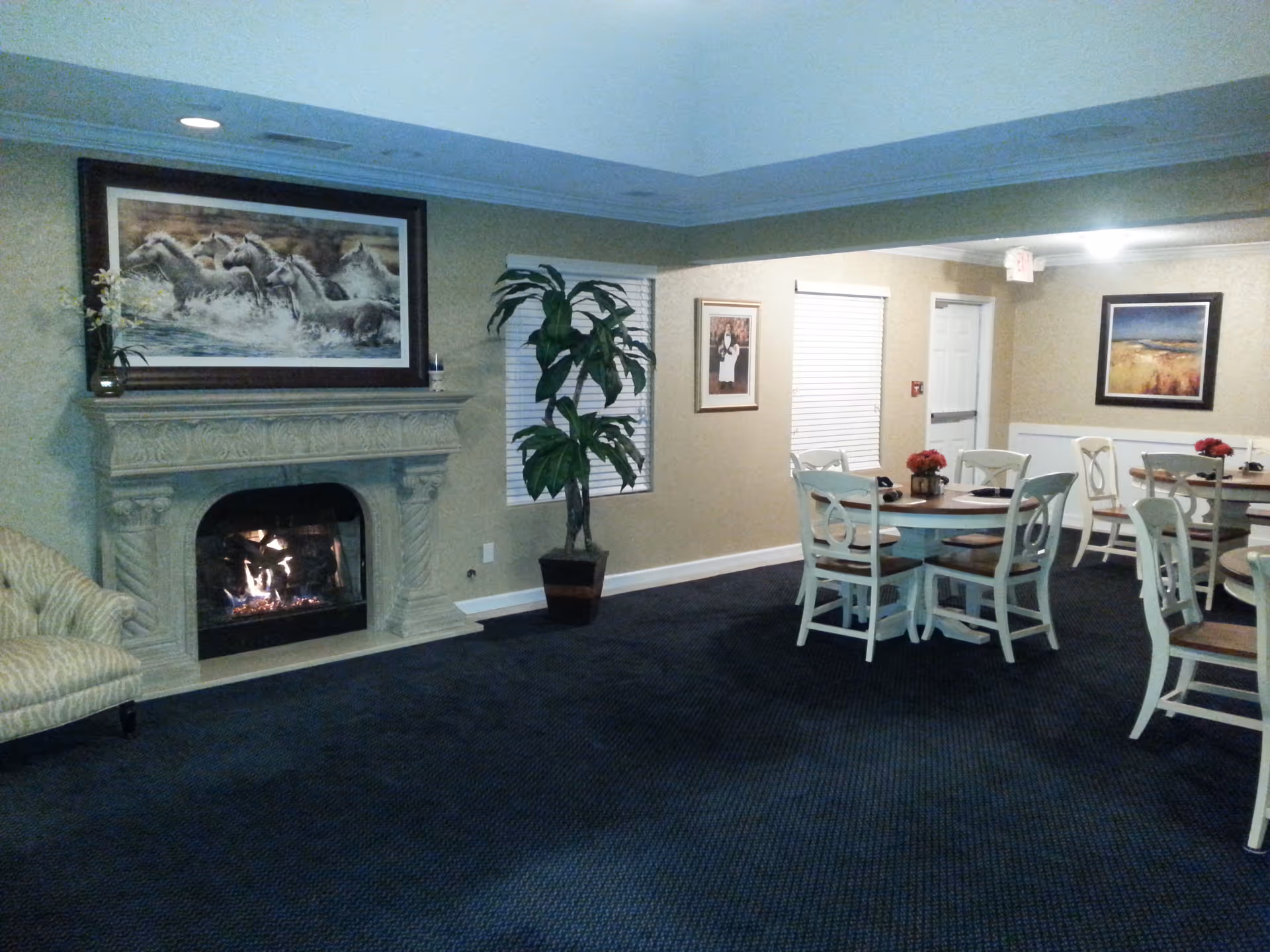 Interior of a senior living facility common area with a lit fireplace, a large framed painting of horses above the fireplace, a potted plant near two windows with blinds, and several round tables with white chairs and red flower centerpieces.