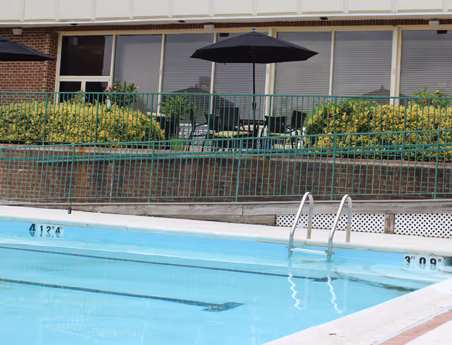 Outdoor swimming pool with clear blue water and metal ladder. Behind the pool, there is a patio area with black patio umbrella, chairs, and tables surrounded by green bushes and a green metal fence. The building exterior with windows and brick walls is visible in the background.