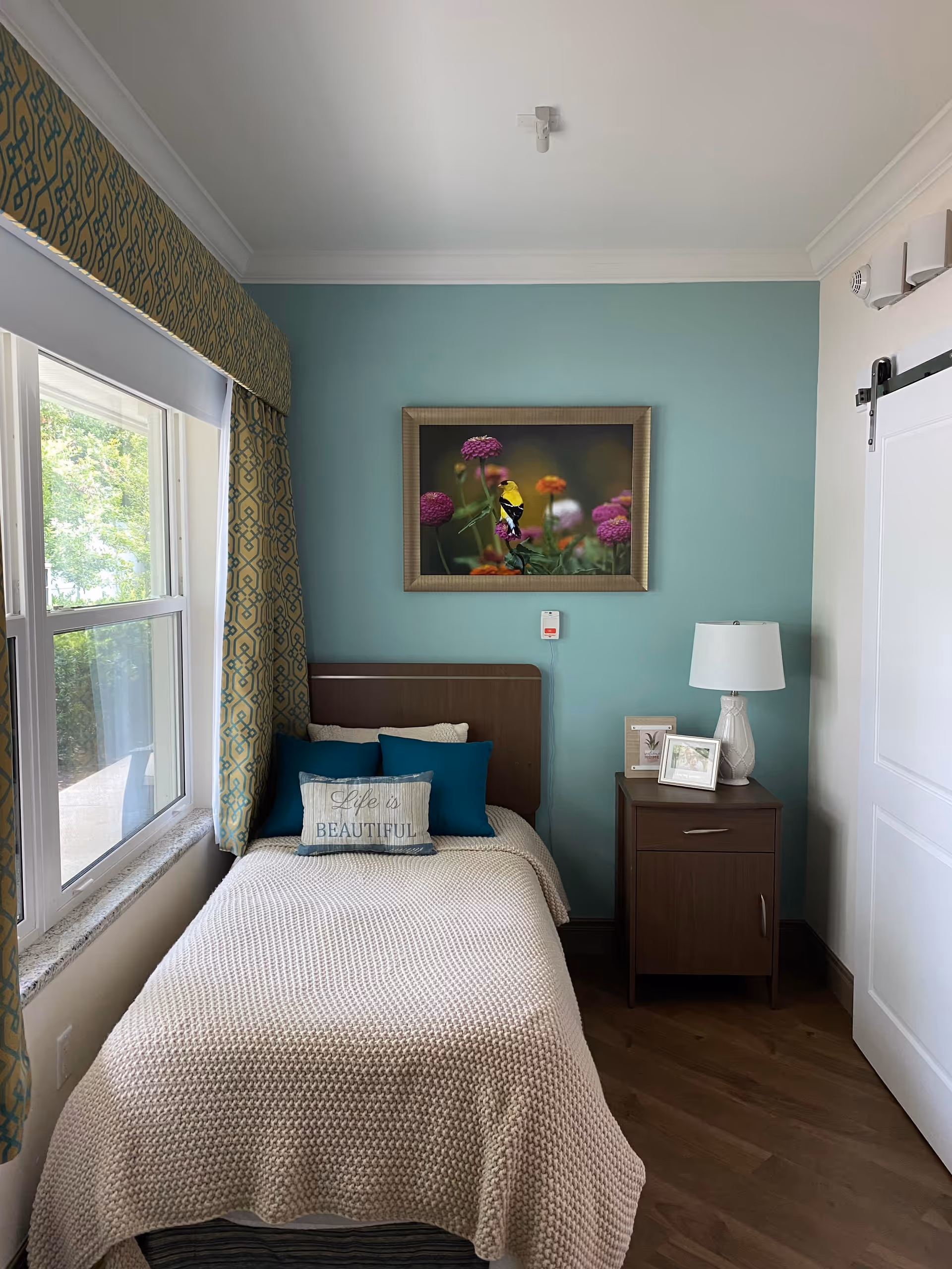 A cozy bedroom with a single bed covered in a beige knitted blanket and decorative pillows, including one that says 'Life is Beautiful'. The bed is positioned next to a window with patterned curtains. A nightstand with a lamp and framed pictures is beside the bed. The wall behind the bed is painted teal and features a framed picture of colorful flowers and a yellow bird.