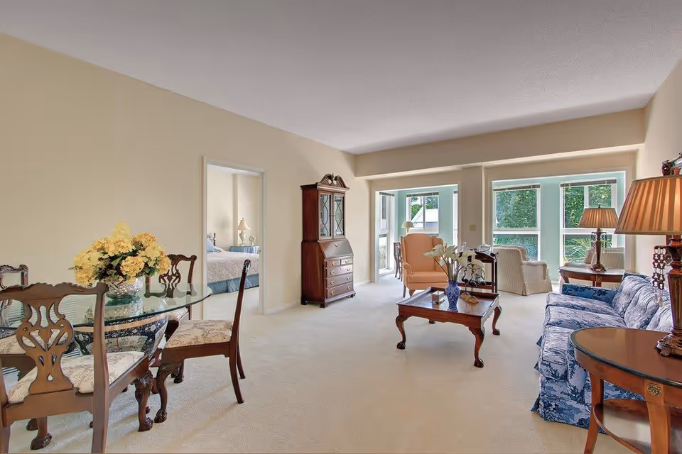 A bright and spacious living room with beige walls and carpet. The room features a blue patterned sofa, a wooden coffee table with a vase of flowers, two armchairs near large windows letting in natural light, a wooden cabinet, and a glass dining table with four wooden chairs. A bedroom is visible through an open doorway.