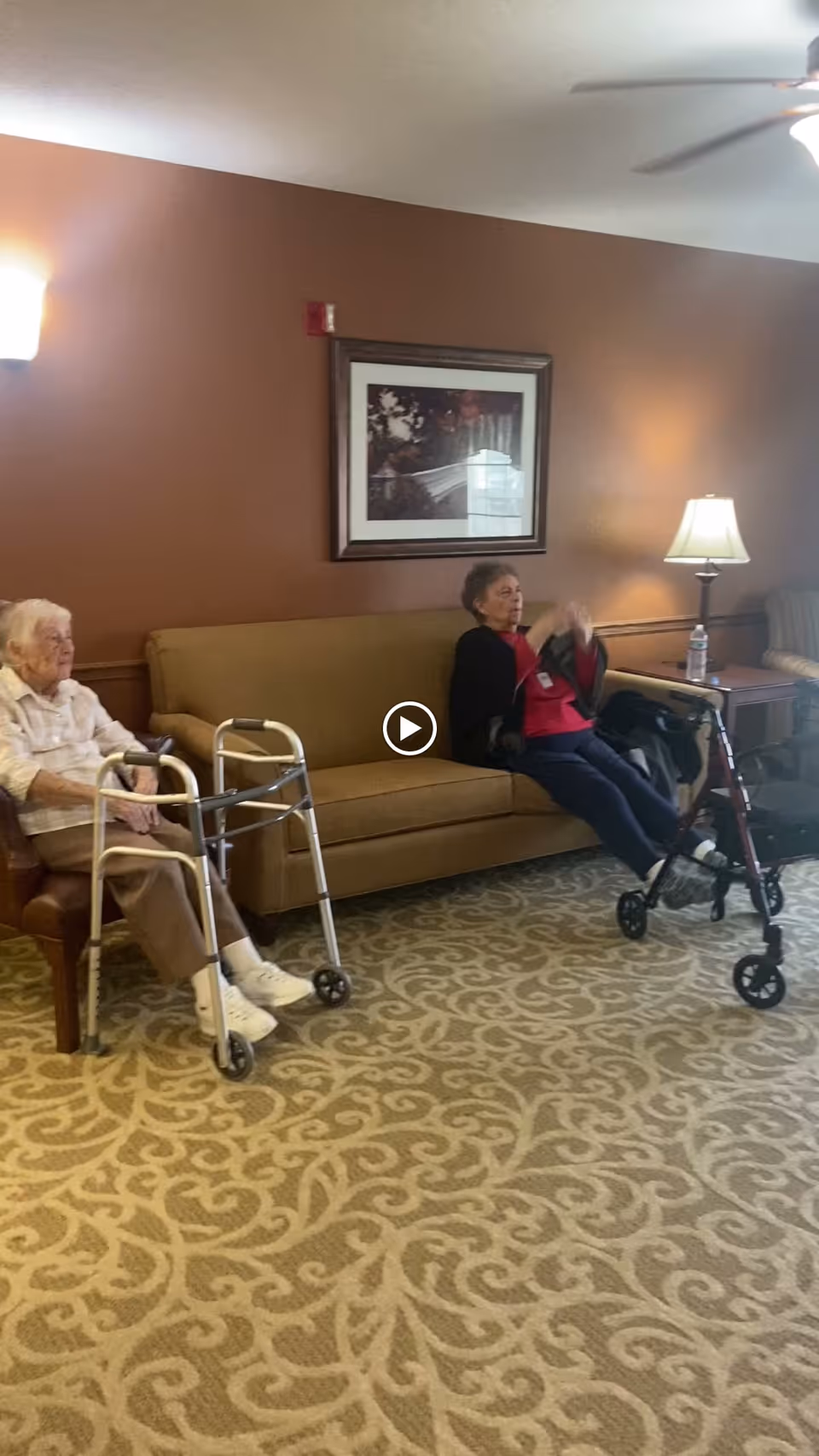 Two elderly women sit in a lounge area with a couch, lamps, framed art, and walkers on patterned carpet.