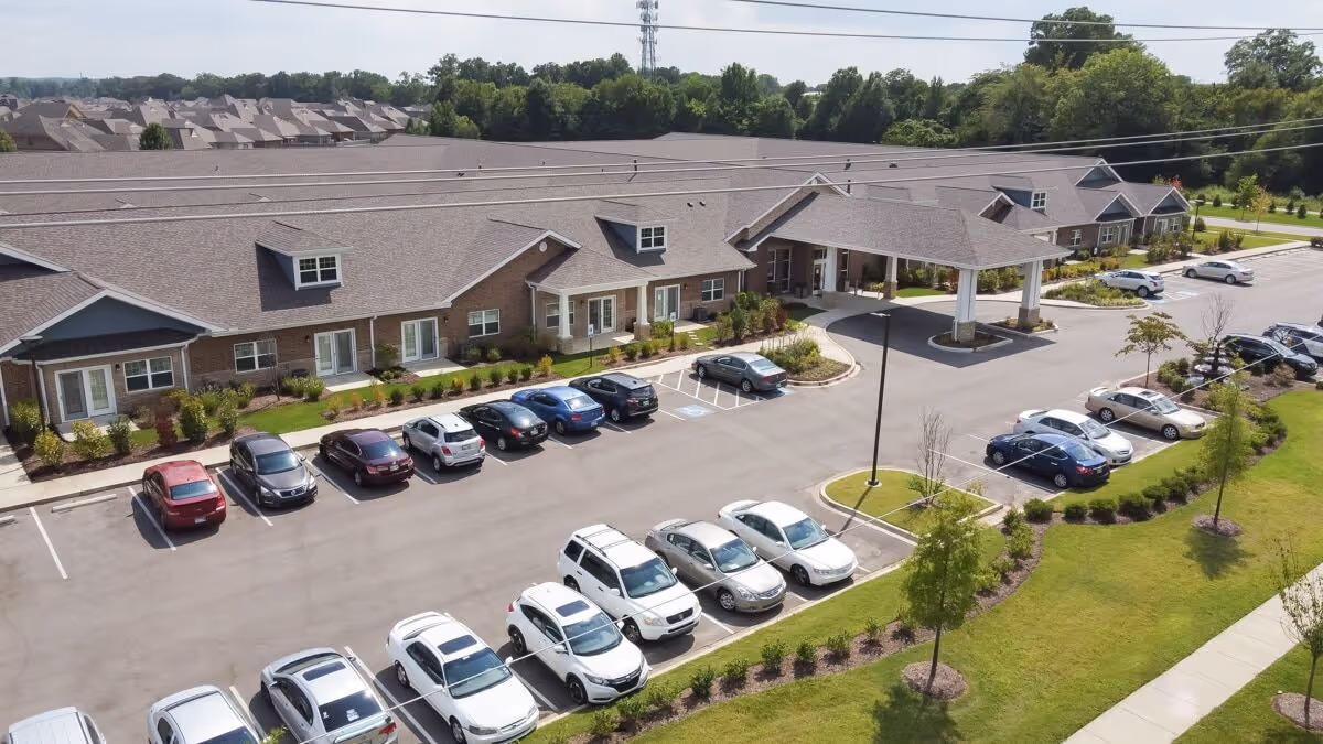 Aerial view of a large senior living facility building with a covered entrance and multiple parked cars in the parking lot. The building has a brown roof and brick exterior, surrounded by landscaped greenery and trees.