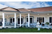 Exterior view of a single-story building with a covered porch supported by white columns. The porch has several white rocking chairs and hanging ceiling fans. In front of the porch is a small lawn with a white decorative fountain.